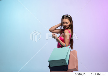 Young woman enjoying successful shopping trip, carrying colorful bags, sunglasses on head, looking at camera on pastel background Young woman enjoying successful shopping trip, carrying colorful bags, sunglasses on head, looking at camera on pastel background 142436162