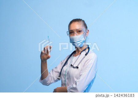 Healthcare worker wearing a protective face mask and safety glasses, holding a syringe with a clear liquid 142436183