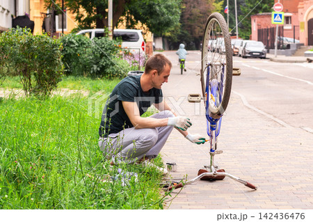 Man examining bicycle wheel near house. Personal vehicle maintenance without service center. Wheel alignment and preparing for upcoming rides Man examining bicycle wheel near house. Personal vehicle maintenance without service center. Wheel alignment and preparing for upcoming rides 142436476