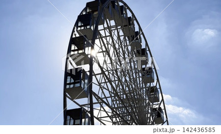 View of a ferris wheel against a clear blue sky with sunlight at background. The sun rays pass through the structure, creating bright highlights and a vibrant summer atmosphere around the attraction View of a ferris wheel against a clear blue sky with sunlight at background. The sun rays pass through the structure, creating bright highlights and a vibrant summer atmosphere around the attraction 142436585