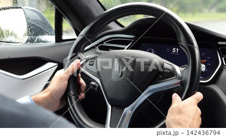Male hands of businessman holds steering wheel while driving electrical vehicle at rain city. Young man commuting to work at rainy day. Businessperson operating electric car on urban road. Close up 142436794
