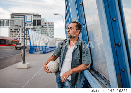 Bearded man with ball under arm using phone while waiting for bus. Bearded man with ball under arm using phone while waiting for bus. 142436821