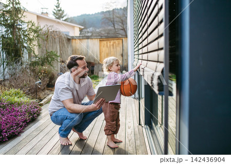 Father controlling smart home exterior blinds using tablet while standing with toddler son. Father controlling smart home exterior blinds using tablet while standing with toddler son. 142436904
