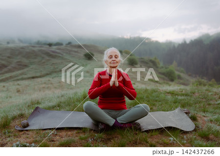 Senior woman meditating in nature on early morning with fog and mountains in background. 142437266