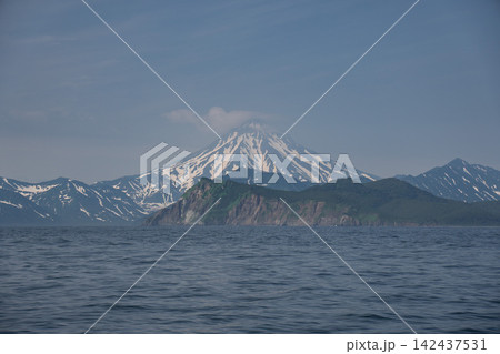 Snowy Vilyuchinsky Volcano rises behind green headland and calm Pacific water in Avacha Bay, Kamchatka, Russia. Nature tourism, volcanic landscape, wilderness, exploration. Snowy Vilyuchinsky Volcano rises behind green headland and calm Pacific water in Avacha Bay, Kamchatka, Russia. Nature tourism, volcanic landscape, wilderness, exploration. 142437531