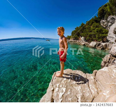 Young boy jumping from coastal rocks into Adriatic Sea on family holiday. 142437683