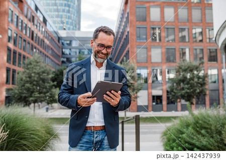 Businessman in front of office building, holding tablet and reading something. 142437939