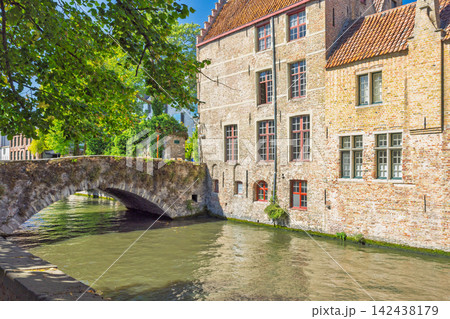Bruges town in Belgium, Europe. Historic Stone Bridge Traverse Calm Canal Waters, Flanked by Traditional Brick Buildings on a Bright Sunny Day, Old Town Architecture Bruges town in Belgium, Europe. Historic Stone Bridge Traverse Calm Canal Waters, Flanked by Traditional Brick Buildings on a Bright Sunny Day, Old Town Architecture 142438179