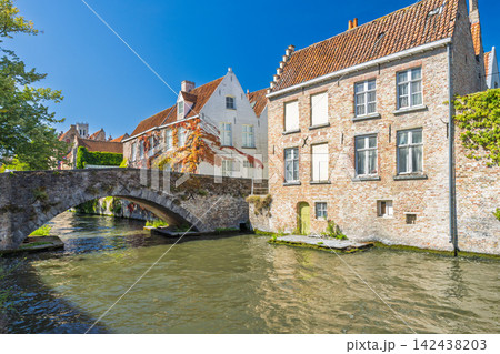 Bruges town in Belgium, Europe. Historic European Canal Scene with Ancient Stone Bridge Arching Over Tranquil Water, Flanked by Traditional Brick Buildings Under Clear Blue Sky Bruges town in Belgium, Europe. Historic European Canal Scene with Ancient Stone Bridge Arching Over Tranquil Water, Flanked by Traditional Brick Buildings Under Clear Blue Sky 142438203