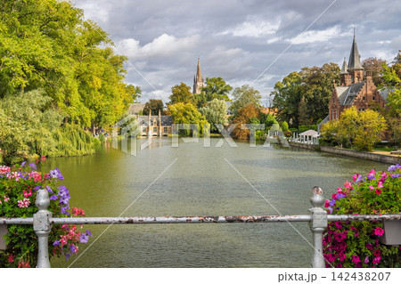 Bruges town in Belgium, Europe. Historic European city lake with lush green trees, blooming flowers, and traditional ancient spires under a dramatic sky 142438207