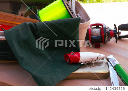 Fishing equipment on a wooden surface. Including a fishing reel with line. A bright green spool of fishing line. A red and white fishing lure. And a pocket knife Fishing equipment on a wooden surface. Including a fishing reel with line. A bright green spool of fishing line. A red and white fishing lure. And a pocket knife 142439326