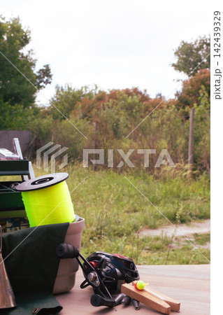Fishing tackle arranged on a wooden surface with a bright yellow line spool, black baitcasting reel, bobbers and weights against a soft green outdoor background, ready for angling 142439329