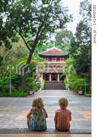 Two children sit on steps facing a traditional Vietnamese temple surrounded by lush tropical greenery in Saigon. Peaceful moment in cultural setting, perfect for family travel, and lifestyle themes. Two children sit on steps facing a traditional Vietnamese temple surrounded by lush tropical greenery in Saigon. Peaceful moment in cultural setting, perfect for family travel, and lifestyle themes. 142440580