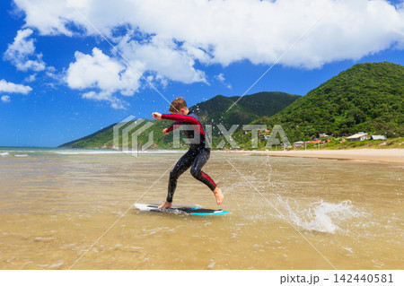 Young boy skimboarding in shallow water on a sunny beach day. Wearing wetsuit, balancing on board with determination and focus. Perfect image for themes of childhood adventure, active lifestyle, Young boy skimboarding in shallow water on a sunny beach day. Wearing wetsuit, balancing on board with determination and focus. Perfect image for themes of childhood adventure, active lifestyle, 142440581