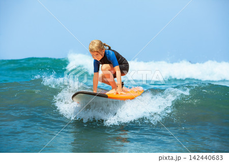 Happy young surfer girl with surfboard have fun on beach, riding on small ocean waves. Summer adventures, active families, parents and kids lifestyle, sports activities on school holiday with kids Happy young surfer girl with surfboard have fun on beach, riding on small ocean waves. Summer adventures, active families, parents and kids lifestyle, sports activities on school holiday with kids 142440683