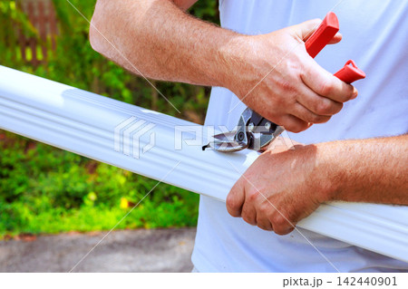 Worker uses sharp shears to cut piece of gutter metal pipe in garden area Worker uses sharp shears to cut piece of gutter metal pipe in garden area 142440901