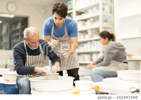 Mature man help to guy working on potters wheel in pottery workshop Mature man help to guy working on potters wheel in pottery workshop 142441348