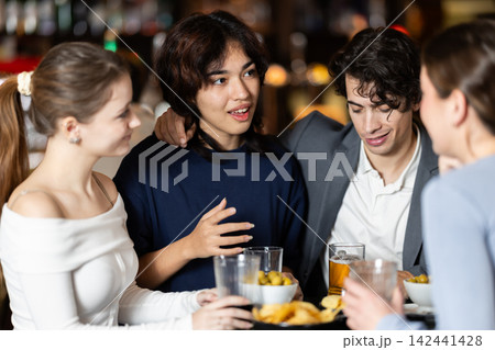 Group of positive young friends talking cheerfully and drinking beer at table in bar 142441428