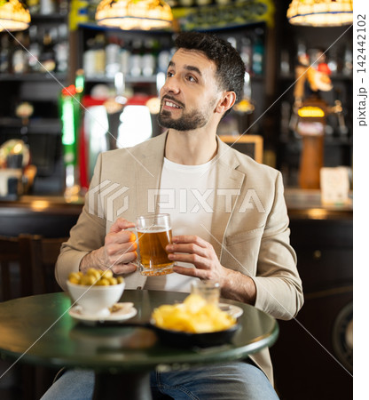 Positive young guy relaxing at table and drinking beer in sports pub, attentively watching match on TV 142442102