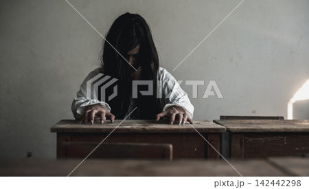 Halloween event features scary woman in ghostly white costume posing menacingly at wooden school desk indoors during creepy holiday celebration in eerie dark classroom setting Halloween event features scary woman in ghostly white costume posing menacingly at wooden school desk indoors during creepy holiday celebration in eerie dark classroom setting 142442298