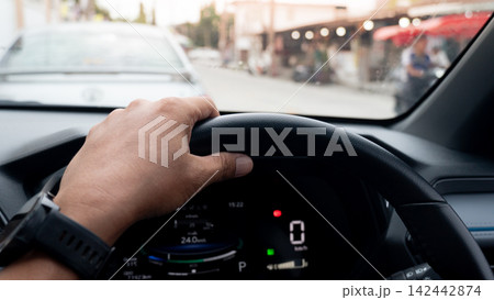 Driver is perspective holding a steering wheel inside a car. Stopped in heavy urban traffic during daytime. Parked behind the sedan in front of the road. Driver is perspective holding a steering wheel inside a car. Stopped in heavy urban traffic during daytime. Parked behind the sedan in front of the road. 142442874