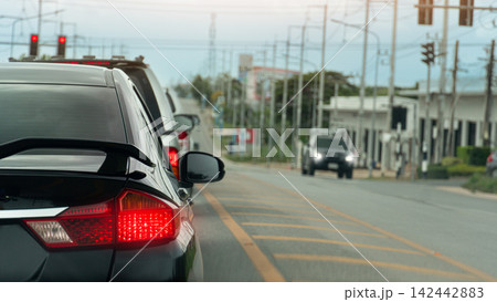 Rear side of black car stop and quere on asphalt road. Car turn on break light. Side road with electric pole and town include trees. on opposite with alone of car driving through. environment at days. Rear side of black car stop and quere on asphalt road. Car turn on break light. Side road with electric pole and town include trees. on opposite with alone of car driving through. environment at days. 142442883