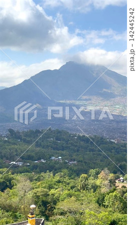 Mountain silhouette above volcanic plain under cloudy blue sky with foreground trees and village, with mountain, volcano, plain, cloud detail, showing sky, peak, silhouette, ridge context 142445242
