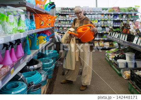 Senior woman shopping for gardening supplies, holding an orange watering can in a store aisle Senior woman shopping for gardening supplies, holding an orange watering can in a store aisle 142448171