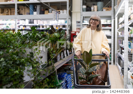 Mature woman smiling, pushing a shopping cart filled with a plant at a nursery Mature woman smiling, pushing a shopping cart filled with a plant at a nursery 142448366