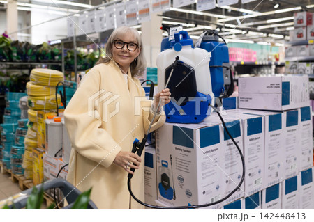 Senior woman in glasses and yellow coat holding a battery sprayer in a hardware store 142448413