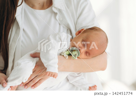Mother holding sleeping infant in white outfit with pacifier, soft natural light illuminating the cozy indoor setting, showcasing a nurturing moment between parent and child Mother holding sleeping infant in white outfit with pacifier, soft natural light illuminating the cozy indoor setting, showcasing a nurturing moment between parent and child 142451774