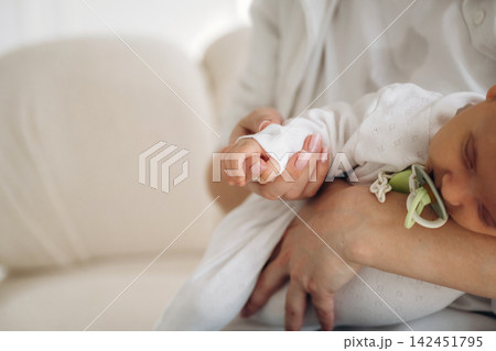 Close-up of a parent holding a sleeping baby in white clothing, with a pacifier attached, seated on a light-colored sofa in a softly lit room Close-up of a parent holding a sleeping baby in white clothing, with a pacifier attached, seated on a light-colored sofa in a softly lit room 142451795