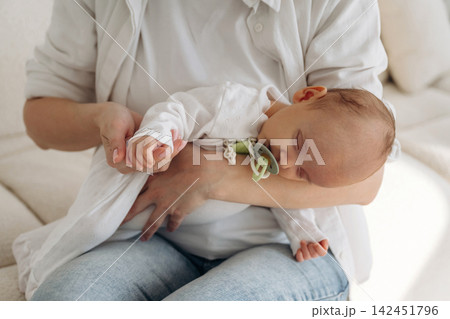 Caucasian woman in white shirt holds sleeping infant in her arms while seated on a light-colored sofa in a cozy indoor setting with soft lighting Caucasian woman in white shirt holds sleeping infant in her arms while seated on a light-colored sofa in a cozy indoor setting with soft lighting 142451796
