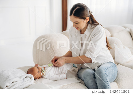 Young woman in white shirt dressing baby boy in cozy living room with soft furnishings and natural light streaming through large windows Young woman in white shirt dressing baby boy in cozy living room with soft furnishings and natural light streaming through large windows 142451801