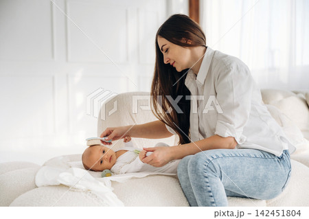 Mother gently brushes hair of her infant child while seated on a soft couch in a bright, airy room with natural light streaming through large windows Mother gently brushes hair of her infant child while seated on a soft couch in a bright, airy room with natural light streaming through large windows 142451804