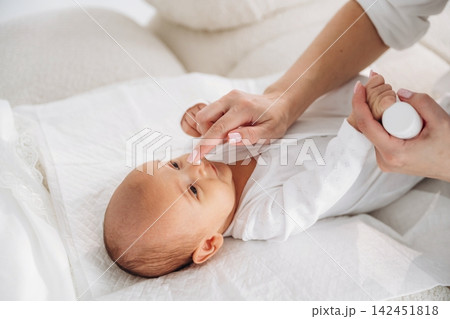 Infant lying on a soft surface while a caregiver applies lotion to the baby's nose, dressed in a white onesie, with a light-colored background and cozy setting Infant lying on a soft surface while a caregiver applies lotion to the baby's nose, dressed in a white onesie, with a light-colored background and cozy setting 142451818