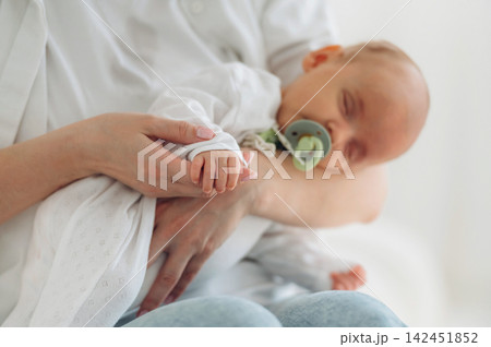 Infant sleeping in caregiver's arms, wearing a white outfit and pacifier, with caregiver's hand gently holding the baby's hand in a bright indoor setting Infant sleeping in caregiver's arms, wearing a white outfit and pacifier, with caregiver's hand gently holding the baby's hand in a bright indoor setting 142451852
