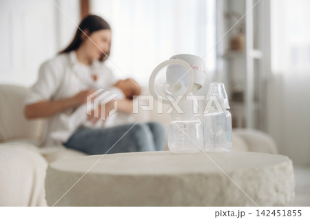 Woman breastfeeding infant while using electric breast pump on a coffee table with baby bottles in a bright, cozy living room setting 142451855