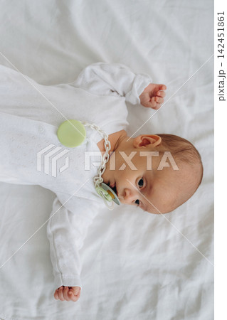 Infant lying on white bedding wearing a white onesie with a pacifier attached to a chain, displaying a calm expression and small hand gestures Infant lying on white bedding wearing a white onesie with a pacifier attached to a chain, displaying a calm expression and small hand gestures 142451861