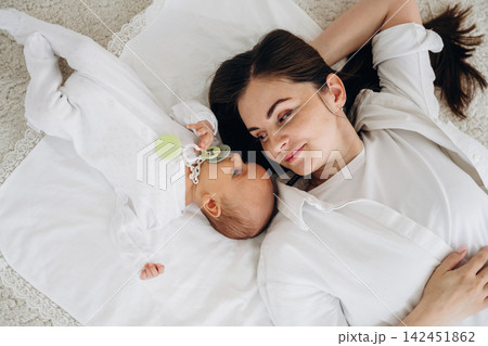 Mother and infant lying on soft blanket, gazing at each other with a pacifier in the baby's hand, bright indoor setting with neutral colors and natural light Mother and infant lying on soft blanket, gazing at each other with a pacifier in the baby's hand, bright indoor setting with neutral colors and natural light 142451862