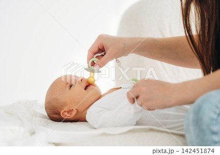Infant lying on a soft surface while a caregiver gently places a pacifier in the baby's mouth, showcasing a nurturing interaction in a bright indoor setting Infant lying on a soft surface while a caregiver gently places a pacifier in the baby's mouth, showcasing a nurturing interaction in a bright indoor setting 142451874
