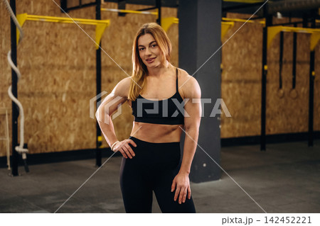 Female athlete with long blonde hair wearing black sportswear stands confidently in a gym with wooden walls and fitness equipment in the background 142452221