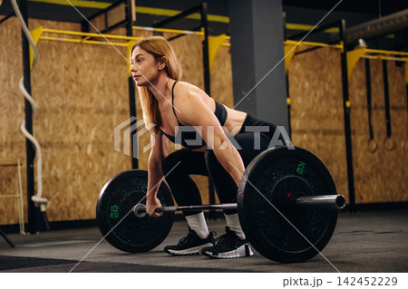 Female weightlifter in black athletic wear prepares to lift a barbell with weights in a gym setting, wooden wall and exercise equipment visible in the background Female weightlifter in black athletic wear prepares to lift a barbell with weights in a gym setting, wooden wall and exercise equipment visible in the background 142452229