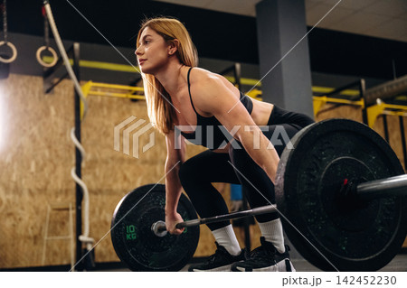 Female athlete performing deadlift with barbell in gym, focused expression, fitness equipment and wooden wall visible in the background Female athlete performing deadlift with barbell in gym, focused expression, fitness equipment and wooden wall visible in the background 142452230