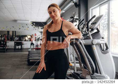 Female fitness enthusiast in black athletic wear poses confidently beside an elliptical machine in a modern gym with mirrors and exercise equipment in the background 142452264
