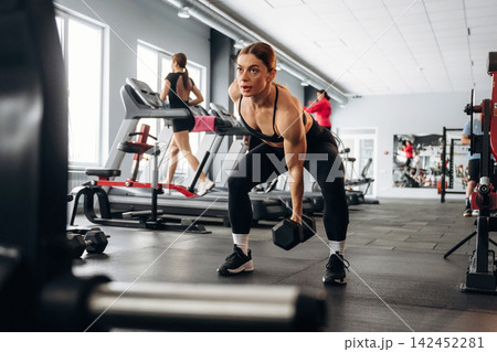 Female athlete performing deadlift exercise with dumbbell in modern gym, fitness equipment and other gym-goers visible in the background Female athlete performing deadlift exercise with dumbbell in modern gym, fitness equipment and other gym-goers visible in the background 142452281