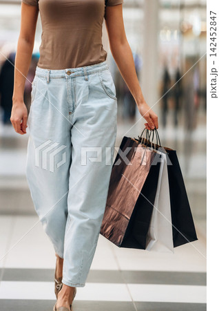 Female shopper wearing casual outfit carries shopping bags while walking through a modern shopping mall with blurred background of other shoppers and stores Female shopper wearing casual outfit carries shopping bags while walking through a modern shopping mall with blurred background of other shoppers and stores 142452847