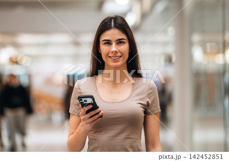 Young woman with long dark hair holding smartphone smiles while standing in a modern shopping mall with blurred shoppers in the background 142452851