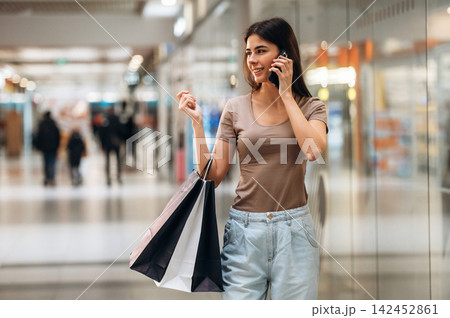 Young woman talking on smartphone while holding shopping bags in a modern shopping mall with blurred shoppers and store displays in the background Young woman talking on smartphone while holding shopping bags in a modern shopping mall with blurred shoppers and store displays in the background 142452861