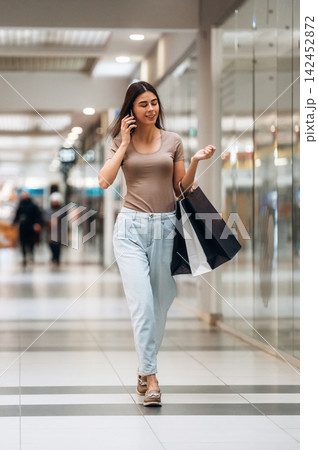 Young woman walking in a shopping mall, talking on her phone while carrying shopping bags, with store reflections visible in the background 142452872